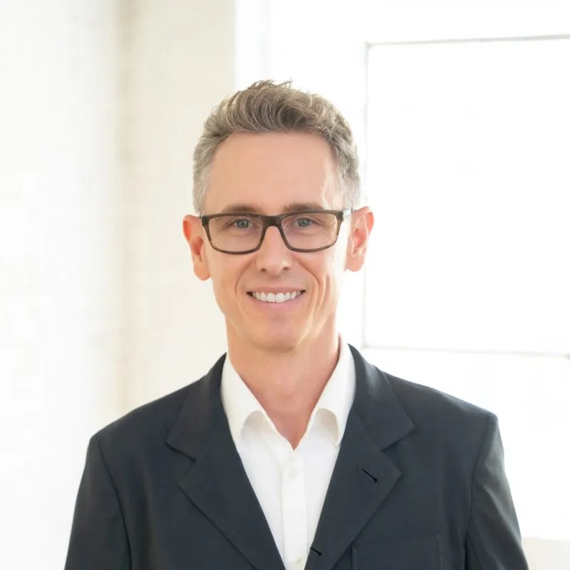 Headshot portrait of Dr. Matthew Alm wearing glasses and a dark suit jacket over a white collared shirt, smiling against a bright background.